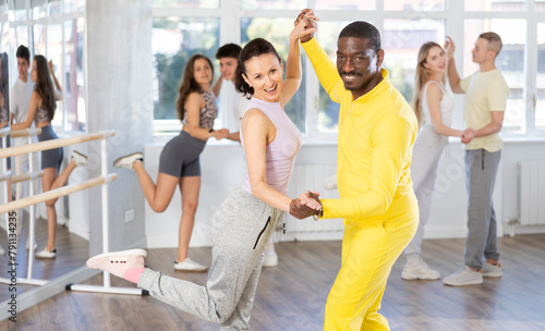 African american man with european girl and group of young active sports people practicing rock and roll dance in modern dance hall