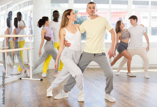 Positive young adult couple, attractive long-haired woman and sporty man dancing playful Latin dance bachata during group class in choreography studio. Active lifestyle concept