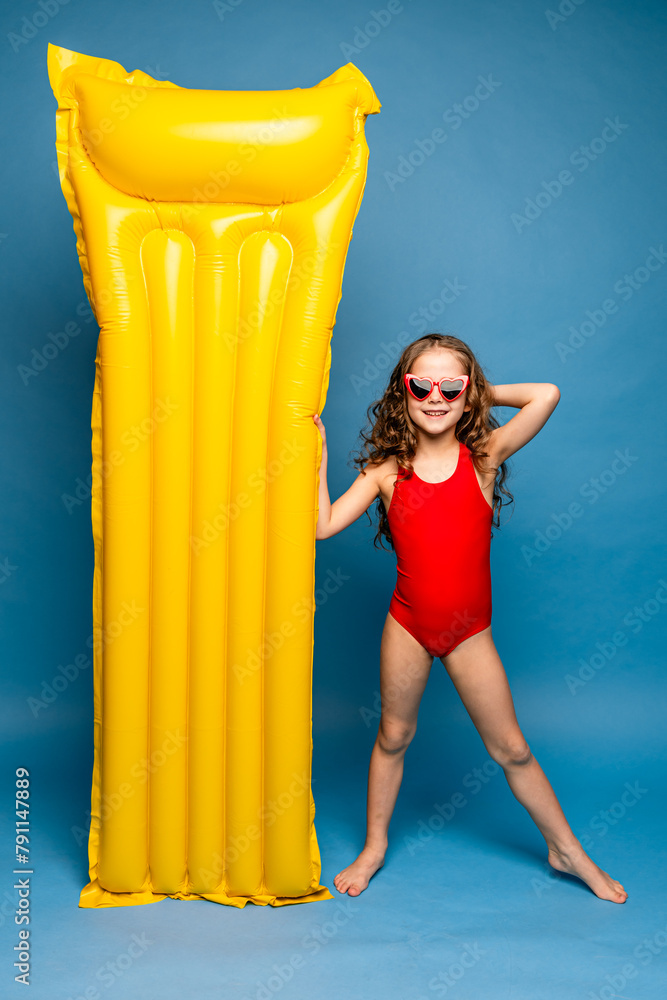 Obraz premium Swimming is fun. Happy little girl wearing red swim suit in sunglasses going to poll with yellow inflatable mattress, blue studio background. Summer vacation concept. Happy family