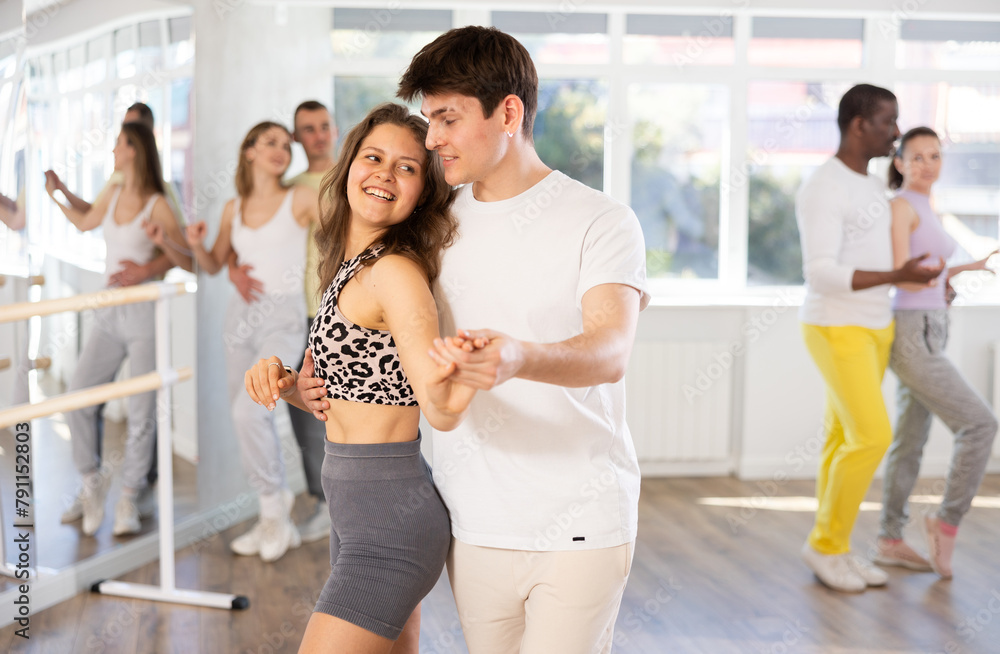 Fototapeta premium Positive young couple, Hispanic guy and fit young girl wearing casual sportswear practicing passionate samba in dance class. Amateur dancing concept..