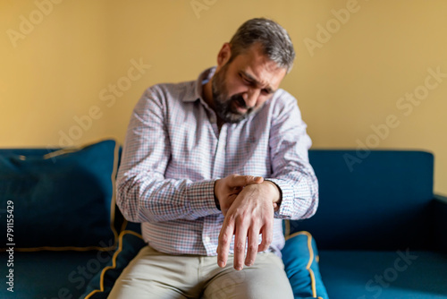 Mature Bearded Man Massaging His Painful Wrist. Man Suffering From Wrist Pain At Home, Sitting on the Sofa