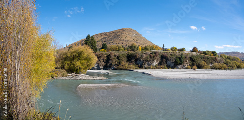 A panoramic view of the Shotover River, clear water flows rapidly over a shallow riverbed with cobblestones, rolling hills and golden autumn trees. A beautiful New Zealand nature landscape. photo