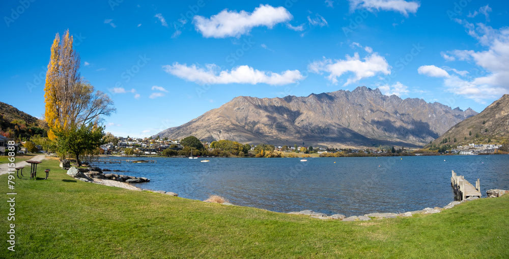 Obraz premium A panoramic view of Lake Wakatipu in Frankton, featuring a lush green grass lawn, a jetty, and the Remarkables mountain range in the background. Queenstown, New Zealand.