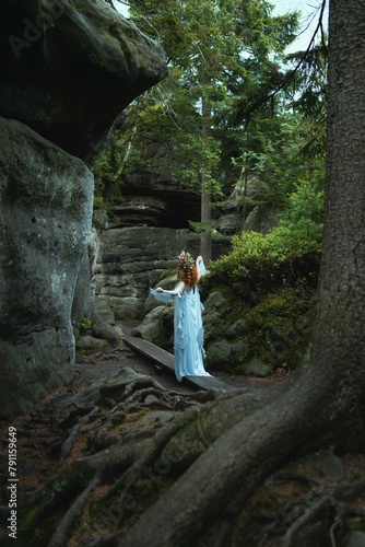 a woman in a fairy blue dress with a flower on her head stands in front of a rock wall.