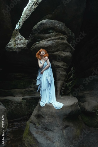 a woman in a fairy blue dress with a flower on her head stands in front of a rock wall.