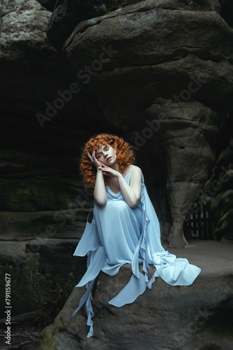 a woman in a blue dress sits in front of a cave with a stone wall behind her.