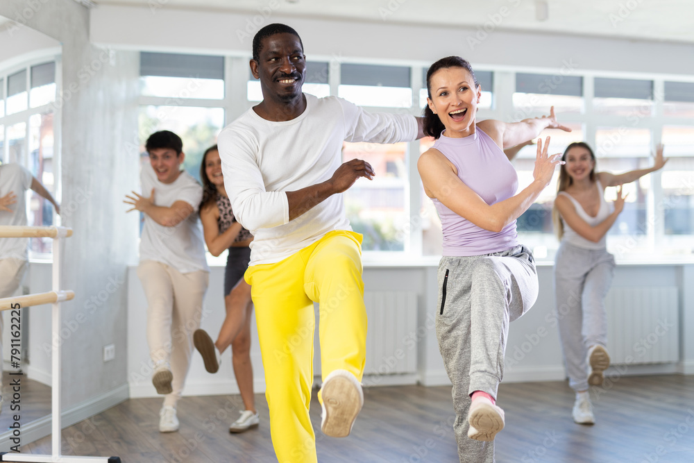 Fototapeta premium African american man with european girl and group of young active sports people practicing rock and roll dance in modern dance hall