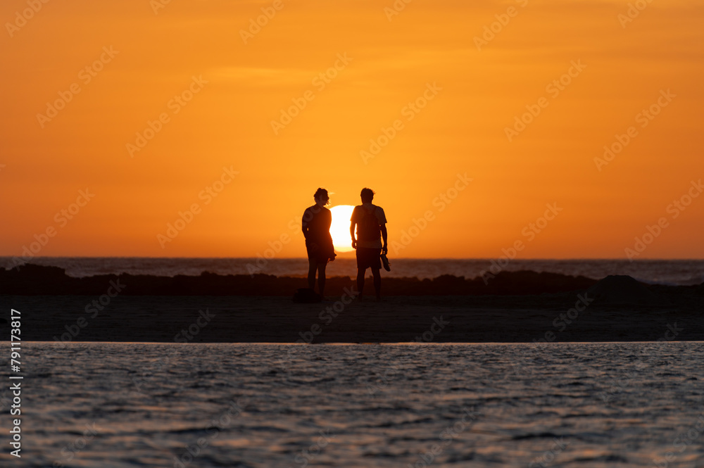 Fototapeta premium Man and woman staying and looking at sunset on ocean beach, orange sky, silhouettes of people on vacation