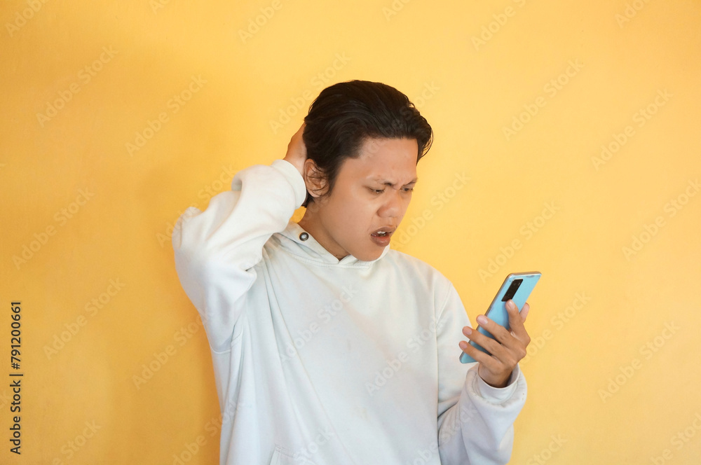 Asian young man showing confused facial expression while holding mobile phone Asian young man showing confused facial expression while holding mobile phone