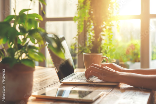 A closeup of hands typing on an open laptop, with coffee and digital tablet beside it on the wooden desk in front of bright window light