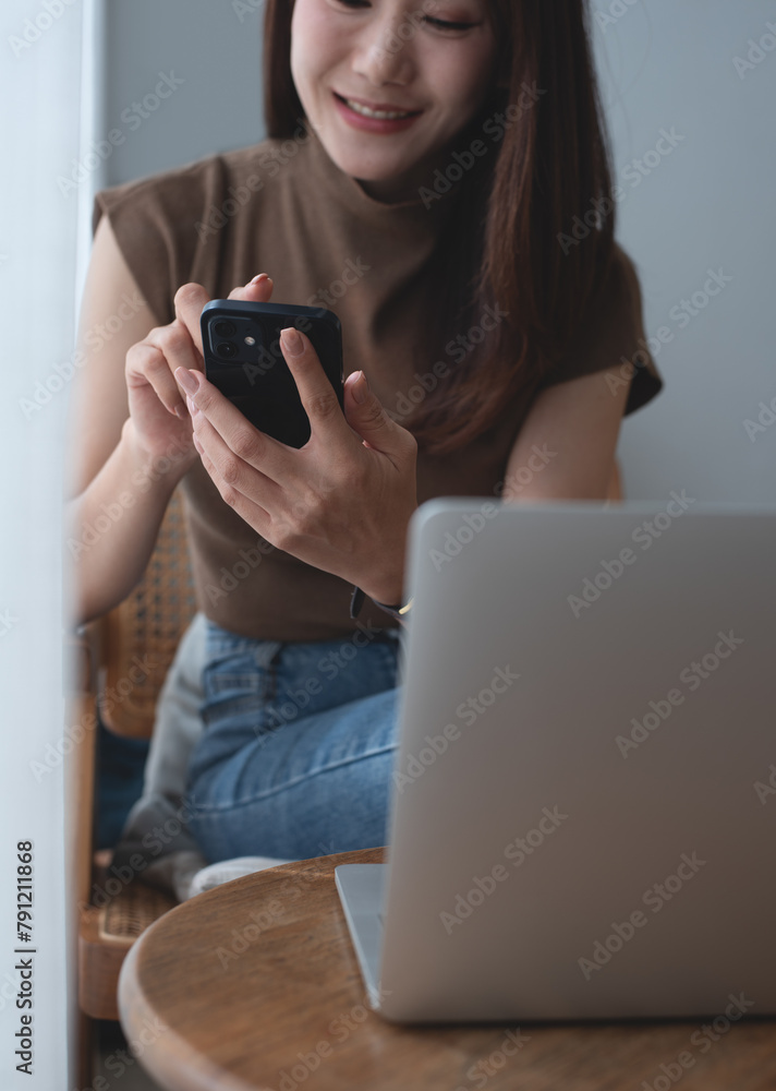 Smiling asian woman in casual wear using mobile phone while working on laptop at coffee shop. Young woman freelancer online working and using smartphone, surfing internet Smiling asian woman in casual wear using mobile phone while working on laptop at coffee shop. Young woman freelancer online working and using smartphone, surfing internet