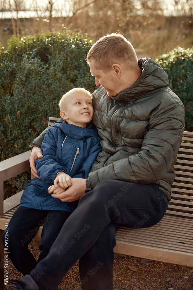 Autumnal Family Affection: Father, 40 Years Old, and Son - Beautiful 8-Year-Old Boy, Seated in the Park.