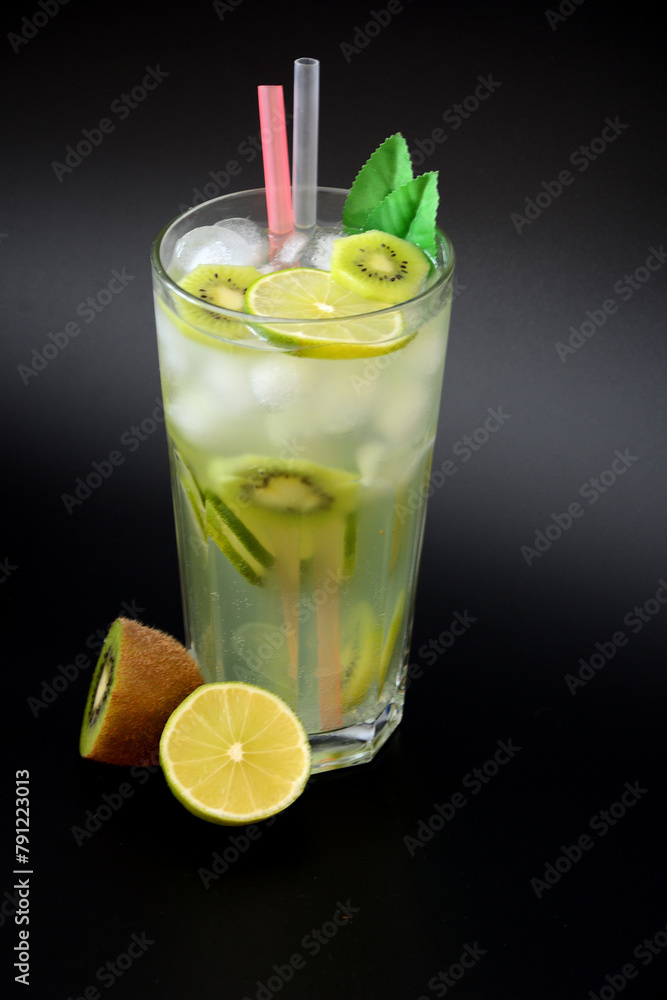 A tall faceted glass of refreshing tropical lemonade with ice and straws on a black background, next to slices of ripe lime and kiwi fruit. A tall faceted glass of refreshing tropical lemonade with ice and straws on a black background, next to slices of ripe lime and kiwi fruit.