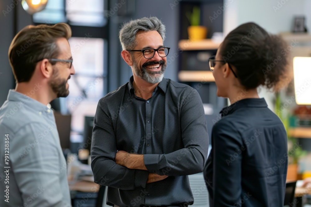 Smiling mature man in eyeglasses looking at camera while standing in office with colleagues Smiling mature man in eyeglasses looking at camera while standing in office with colleagues