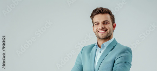 A young businessman in a light cyan suit stands with his arms crossed, smiling against a white backdrop.