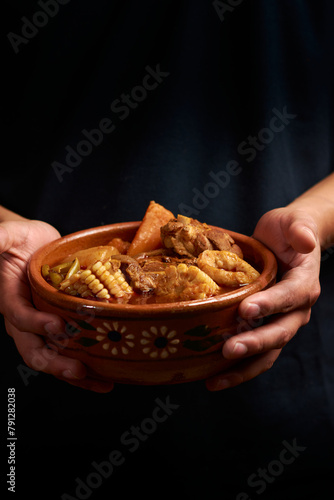 Chilatole with pork, vegetables, carrot, green beans, potato, corn and ears of dough, the clay plate is held by two hands, black background, human part, hands, plate held by hands,