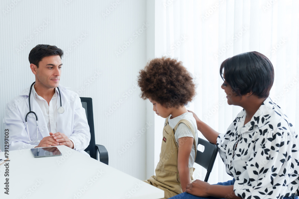 Attentive male doctor in white coat communicates with young patient, mother beside, in a pediatric office, reflecting a calming medical dialogue. Young medic consults with child patient Attentive male doctor in white coat communicates with young patient, mother beside, in a pediatric office, reflecting a calming medical dialogue. Young medic consults with child patient