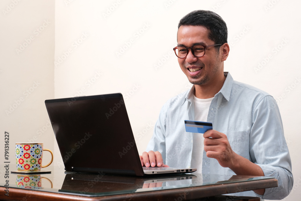 A man smiling happy in front of a laptop with hand holding credit card A man smiling happy in front of a laptop with hand holding credit card