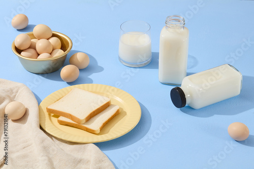 A yellow dish featured two slices of sandwich arranged with a golden bowl containing eggs. Milk is filled inside a cup and bottles. An beige apron is featured on blue surface