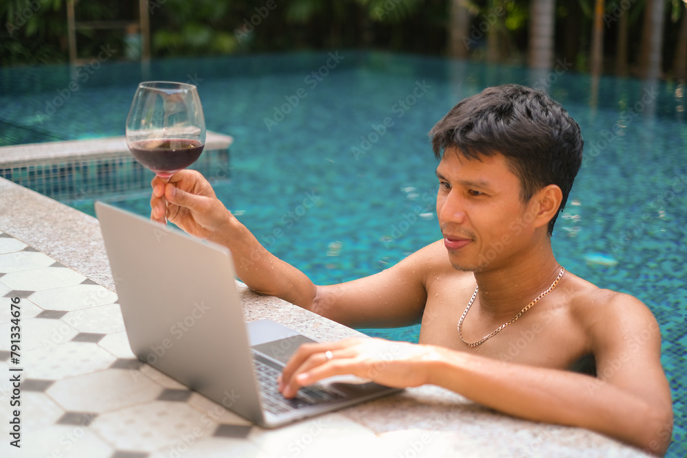 Asian man with a glass of red wine looking happy while checking mail, news, social networks, working on notebook, writing blog, studying at home, resort, hotel on weekend. Online freelance concept. Asian man with a glass of red wine looking happy while checking mail, news, social networks, working on notebook, writing blog, studying at home, resort, hotel on weekend. Online freelance concept.