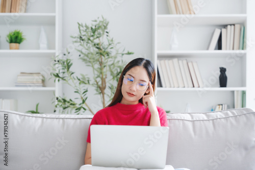 Happy young Asian woman wearing eyeglasses using laptop while seated on couch at home