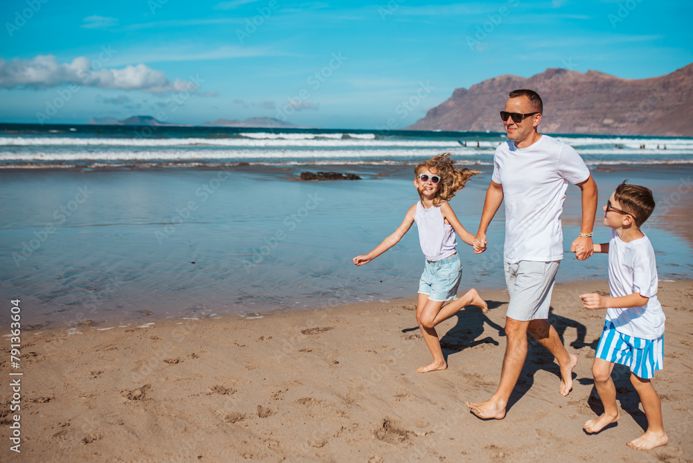 Father with two kids running on beach. Young family enjoying sandy beach, looking at crystalline sea in Canary Islands. Concept of beach summer vacation with kids. Father with two kids running on beach. Young family enjoying sandy beach, looking at crystalline sea in Canary Islands. Concept of beach summer vacation with kids.