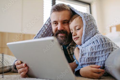 Little boy watching cartoon movie on tablet with father, lying under blanket on floor in kids room. Dad explaining technology to son, digital literacy for kids. © Halfpoint