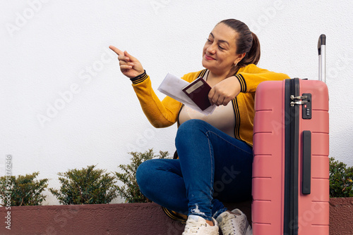 Happy Latina woman with her pink suitcase going on vacation. Excited woman ready to travel and enjoy the summer. Girl looking at her tickets and pointing where to go.