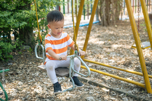 Cute smiling little toddler kindergarten boy child having fun swinging at children playground in summer sunny day
