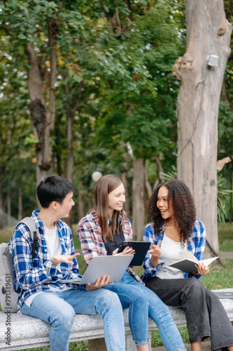 Three young college students is reading a book while relaxing sitting on grass in a campus park with her friends.