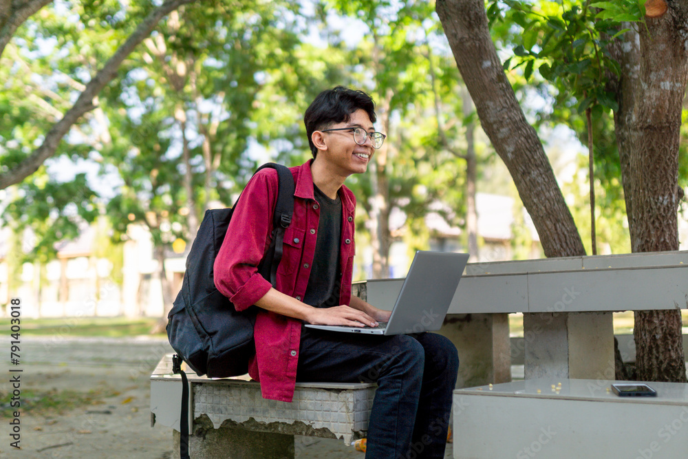 Fototapeta premium Asian college student sitting and focused to his laptop screen. Asian young male sitting outdoors using laptop computer, typing, working freelance project