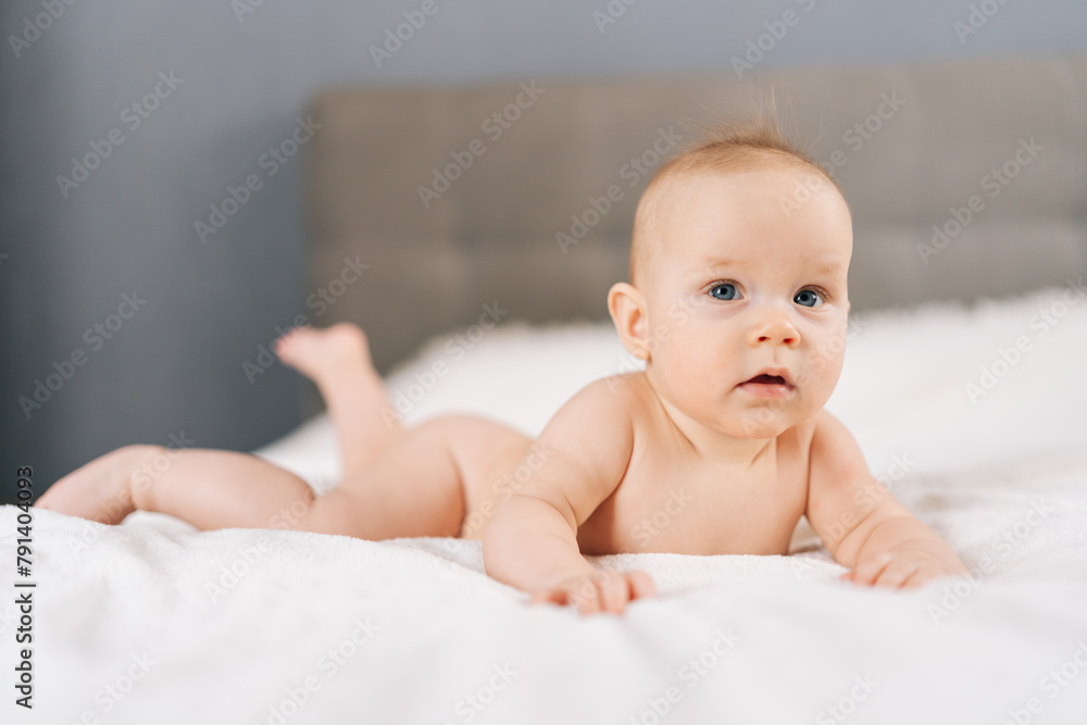 Portrait of happily adorable infant baby lying on bedroom bed, enjoying morning relaxation indoor at home, looking at camera. Carefree healthy babyhood, healthcare and pediatrics care concept