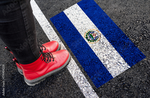 a woman with a boots standing on asphalt next to flag of El Salvador and border
