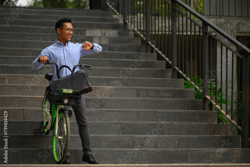 Smiling businessman with his bicycle in front of the building and checking time on wristwatch