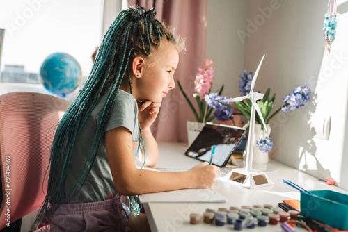 Girl with dyed long braided hair writing in book by wind turbine model at table photo