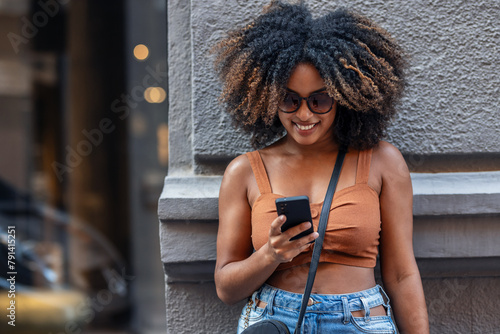 Happy woman using smartphone while carrying shopping bags leaning against the wall in the city