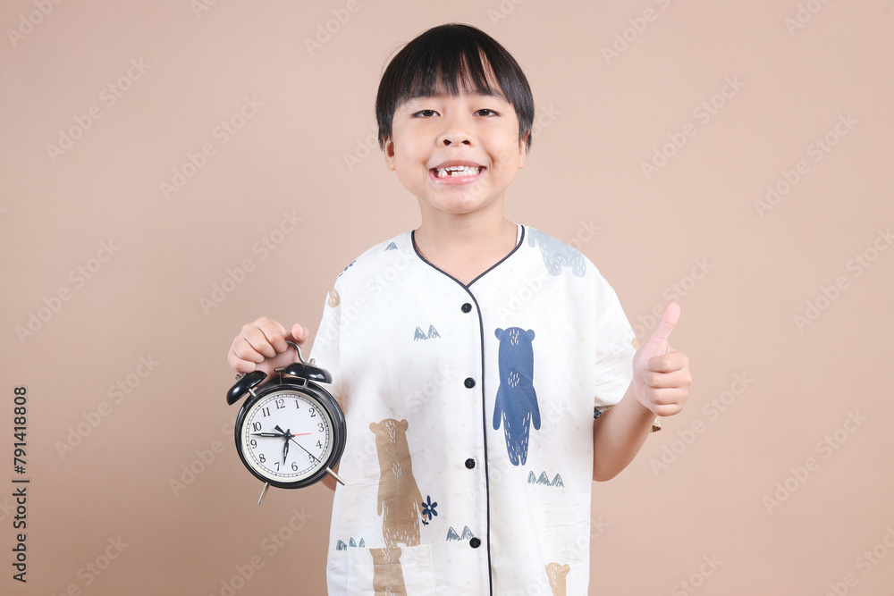 Young Boy In Pajamas Showing Alarm Clock While Showing Thumb Up Isolated on Beige Background Young Boy In Pajamas Showing Alarm Clock While Showing Thumb Up Isolated on Beige Background