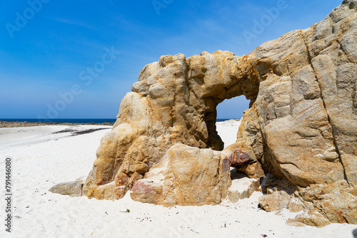 Plage de sable blanc bordée par une arche naturelle creusée dans la roche, baignée par des eaux turquoises, sous un ciel bleu en Finistère Nord, Bretagne.