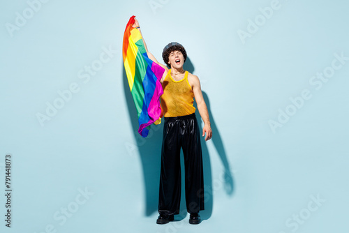 Full body young happy gay Latin man wears mesh tank top hat clothes hold in hand striped rainbow flag isolated on plain pastel light blue cyan background studio Pride day June month love LGBT concept.