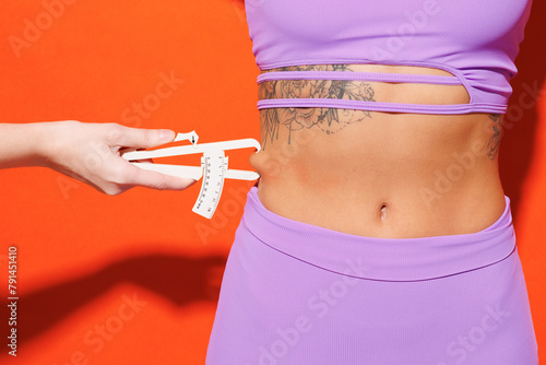 Close up cropped young fitness trainer instructor sporty woman sportsman in purple top clothes in home gym hold use caliper for waist isolated on plain orange background Workout sport fit abs concept.