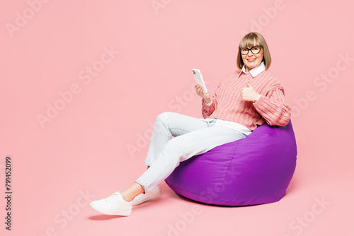 Full body elderly woman 50s years old wear sweater shirt casual clothes glasses sit in bag chair hold use mobile cell phone show thumb up isolated on plain pink background studio. Lifestyle concept.
