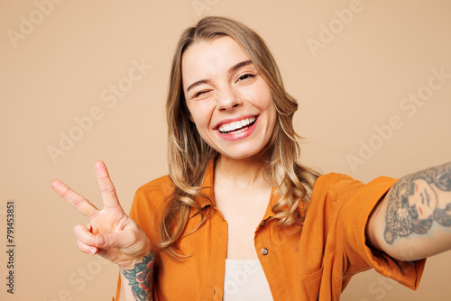 Close up young fun woman she wear orange shirt casual clothes doing selfie shot pov on mobile cell phone show v-sign isolated on plain pastel light beige background studio portrait. Lifestyle concept. © ViDi Studio