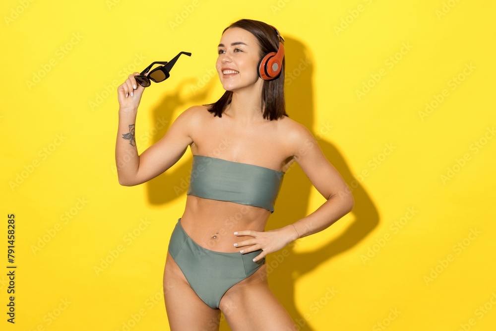 This photo depicts a young woman in a stunning green swimsuit on a yellow background and listening to music through her headphones This photo depicts a young woman in a stunning green swimsuit on a yellow background and listening to music through her headphones