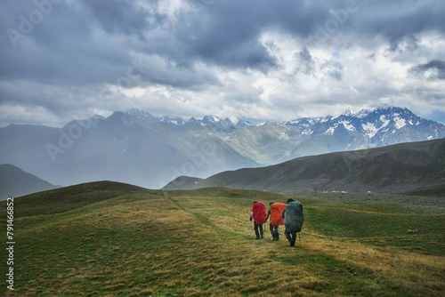 Three hikers with backpacks trek along a grassy ridge against a backdrop of dramatic mountain peaks and cloudy skies