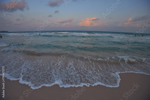 Sun sets, creating a pastel sky, beachgoers bask in the tranquil ocean waves