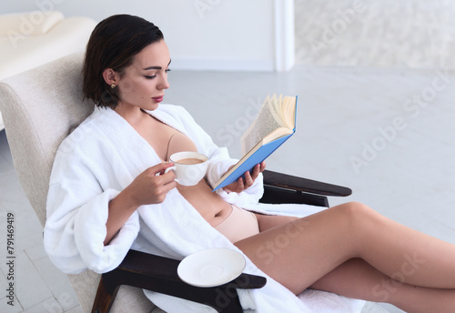 Cute girl in bathrobe sits on armchair, drinks coffee and reads book. Portrait of beautiful young girl at home in the morning
