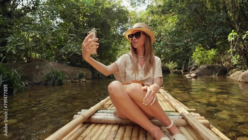 Female tourist floats on bamboo raft. Woman tourist enjoying the bamboo rafting on the river, films beautiful nature landscape on smartphone. Vacation, tropical tourism, travel park adventure concept. photo