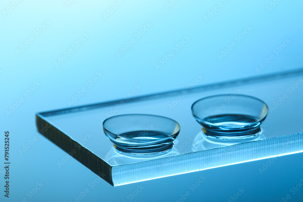 Fototapeta premium Pair of contact lenses on glass against light blue background, closeup