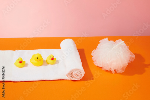 A bath white towel is spread out on the orange table with several size of rubber duck toys placed above, next to a bath sponge. Photo with space for advertising or designing from front view