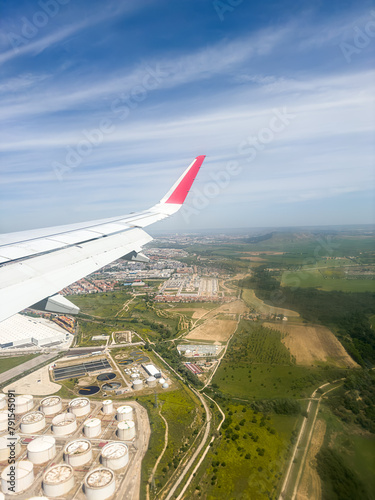 A white wing of an aircraft, adorned with a crimson streak, gliding above a verdant rural scenery teeming with trees, either climbing or descending